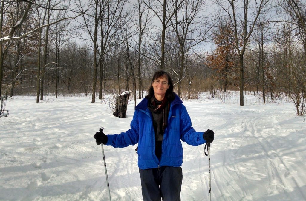 Woman in a blue snow jacket standing in the snowy woods with ski polls