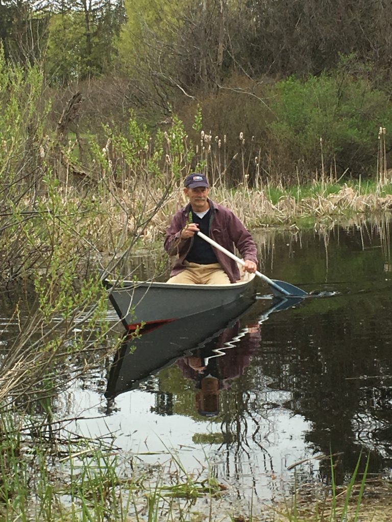 Man with a mustache and baseball cap floating in a small paddle boat on a pond with cattails.