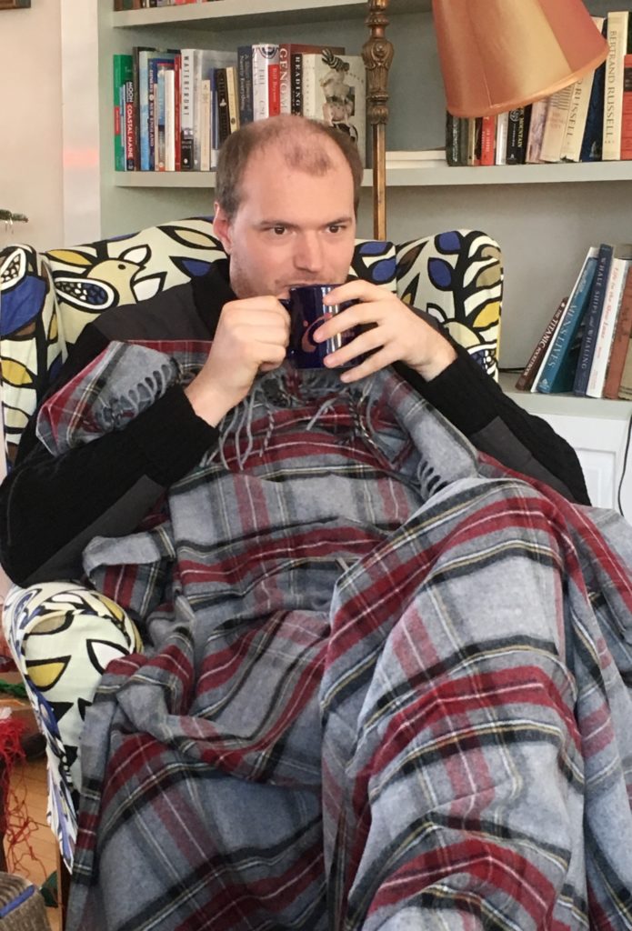 A man sits with a cup of coffee beside a book case covered in a red and black tartan blanket enjoying some coffee.