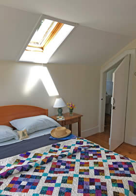 Vaulted room with beige walls, wide plank wood floors, a sunny skylight window and bed with colorful quilt