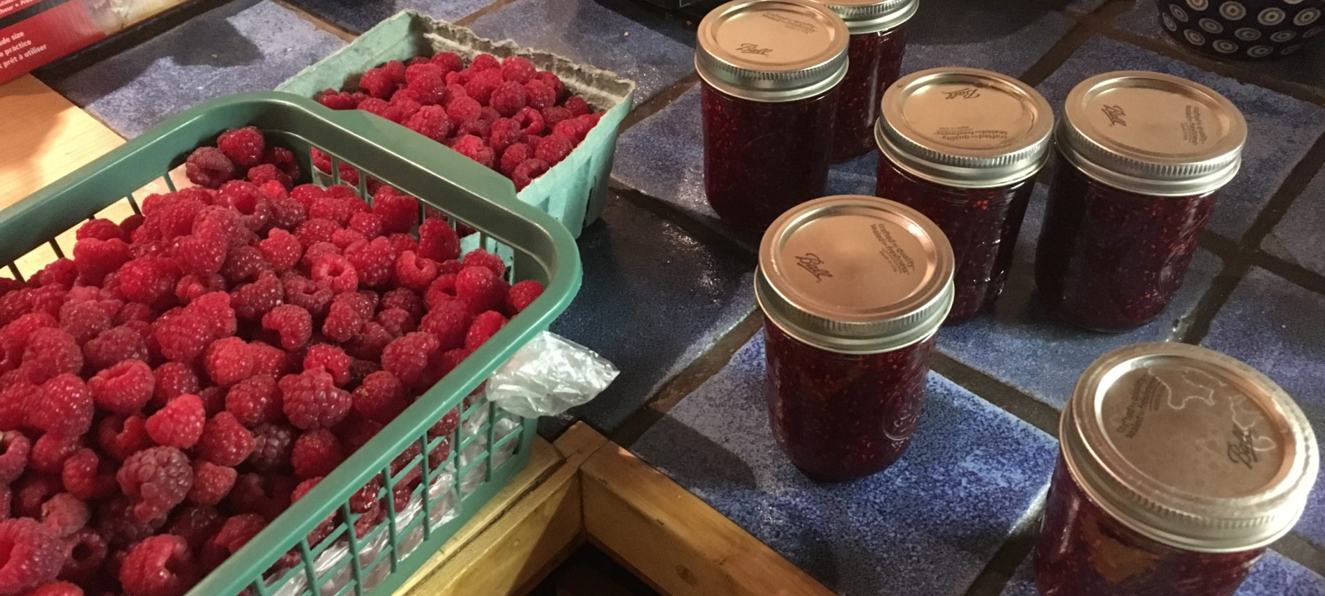 A green basket of raspberries beside jars of raspberry jam.