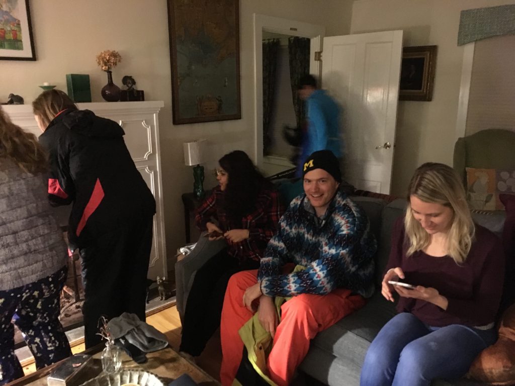 A young man with orange snow pants on looks at the camera smiling, one of a group of 6 young adults in winter clothes warming themselves by an indoor fire place.