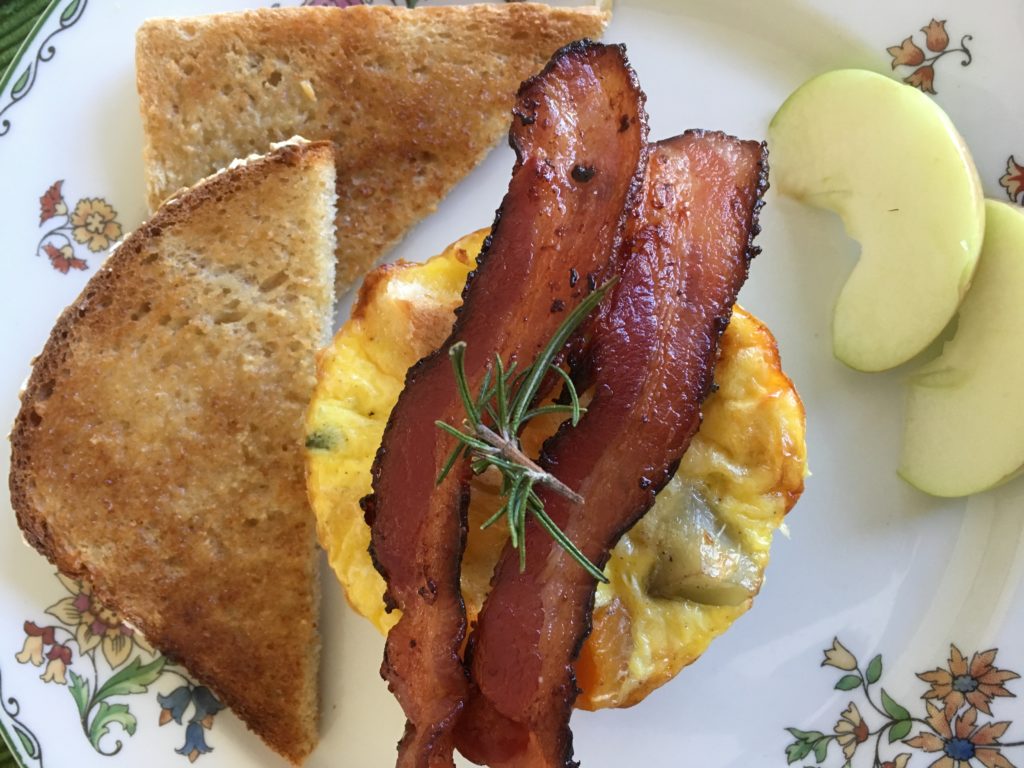 Two strips of bacon laid across egg strata with toast and apple slices on a white china plate.