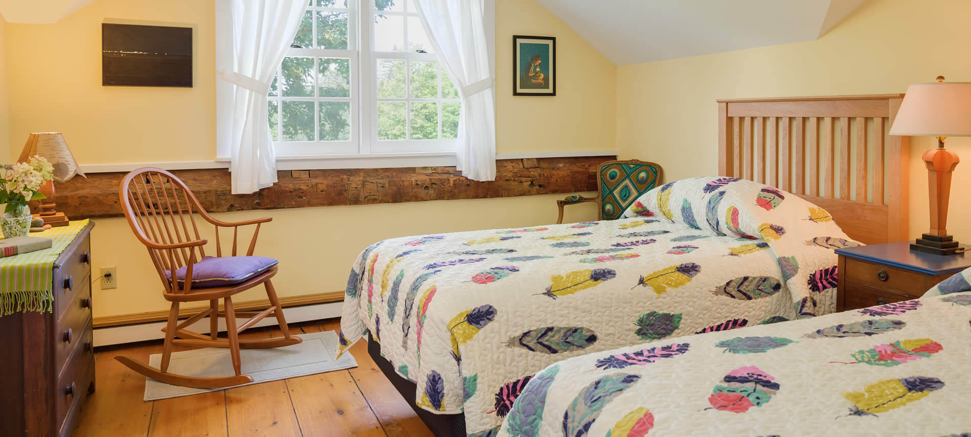 Bright vaulted room with yellow walls, wide plank wood floors, wood rocking chair and two beds with colorful quilts